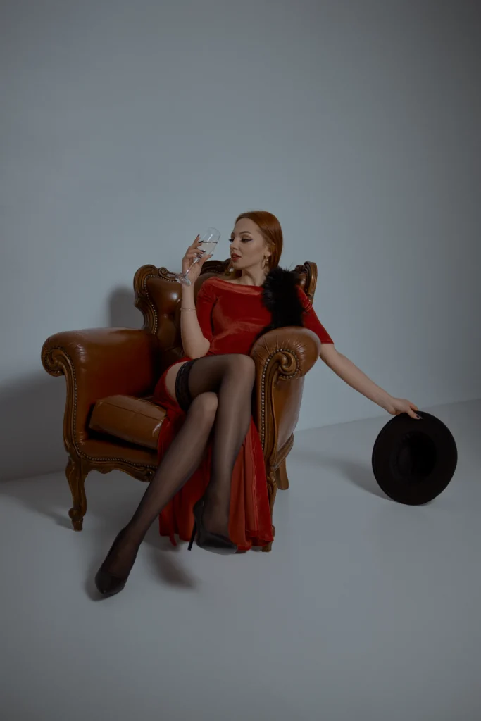 дівчина в панчохах та червоному платті у сидить в кліслі та п'є шампанське